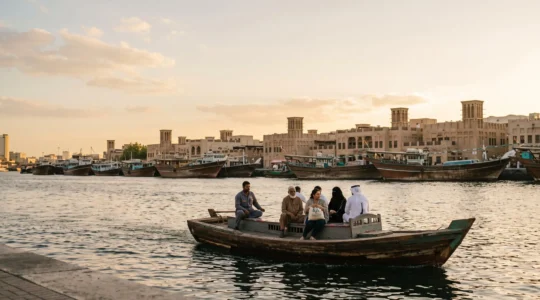 Un abra traditionnel en bois traverse le Dubai Creek au coucher du soleil, avec les dhows de Deira en arrière-plan